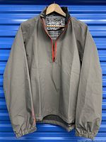 Front view of gray half-zip shell jacket with red zipper and elastic cuffs hanging on wooden hanger.