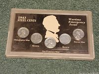 Front side of the hard plastic case containing five 1943 steel cents on display board with Lincoln silhouette and mint names.