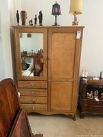 Front view of light wood armoire with a mirror on the left door, three drawers at the bottom, and a solid right door. Small wooden knobs on drawers and doors. Displayed with decorative items on top and chair partially visible beside.