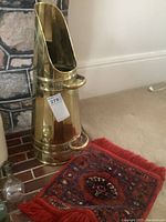 Brass coal scuttle displayed next to stone fireplace, showing polished surface, tall ovoid shape with double handles and open top.