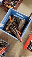 Blue plastic crate filled with assorted hand tools including chisels, files, and sharpening stones, showing wooden handles and metal surfaces.