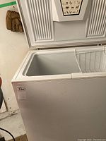 Closed view of white Kenmore mini freezer with an open hinged lid, showing storage basket inside and part of the wall and floor around it.