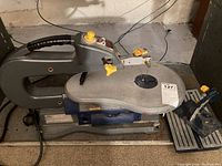 View of entire scroll saw showing metal frame, work surface with blade, foot pedal, power cord and yellow control knobs.