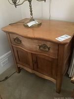 Front perspective of the antique small wooden washstand showing curved edges, single drawer with brass handles, and double door cabinet base.