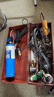 View inside metal toolbox showing blue handheld propane torch, soldering irons with cords, wooden-handled torch, and multiple soldering accessories.