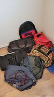Photo of nine assorted bags arranged on the floor, showing variety of styles and colors, mainly duffle bags and one backpack.