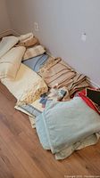 Photo showing blankets folded and stacked on floor, multiple fabrics and colors including light cream and blue.
