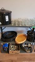 Front view of shelving unit with large black enamel canning pot on top shelf with many mason jars arranged next to it. Below are two black enamel roasting pans, wicker basket bowl, metal colander, and box of metal canning rings and lids.