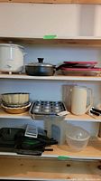 White Proctor Silex slow cooker with black lid, various bake pans including metal muffin tray, ceramic dish, juice jugs, glass bowl, and plastic containers on wooden shelves