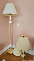 Photo showing full view of brass floor lamp with white cream shade, brass table lamp with cream shade beside it, and extra lamp shade placed in front.