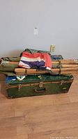 Photo showing green metal military trunk with brass latches, wood frame canvas cot, folded Union Jack flag, and camouflage tarps on top