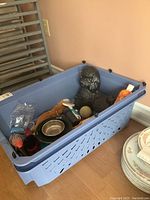 Photo of blue plastic pet carrier containing ceramic poodle figurine, grooming brushes, pet food dishes, ball, and toys.