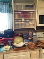 Photo of kitchen cabinet shelves showing organized plastic storage containers with lids, glass jars, and some mixing bowls below on a countertop. Also visible are wicker baskets, and insulated lunch bags in the foreground.