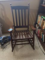 Front view of dark brown painted wooden rocking chair with vertical slats, armrests, and visible chips on seat.