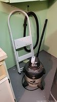 Overall lot showing the shop vac and folding ladder placed in a corner with a green wall.