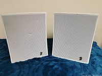 Front view of two white Infinity Outrigger Jr. speakers with curved grills, placed side by side on blue cloth.