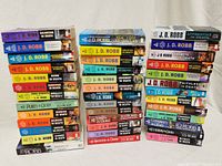 Three vertical stacks of paperback books showing spines of predominantly J.D. Robb titles with some bonus books by Nora Roberts and others. Titles and condition visible.