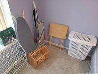 Photo of corner showing ironing board, Oreck upright vacuum, wooden TV tray, white plastic hamper, and white wire shoe rack.