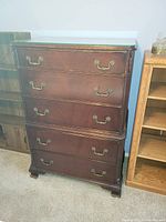 Photo showing a dark wood 5 drawer dresser with brass handles and decorative carving.