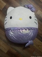 Large Hello Kitty Squishmallow plush toy on wooden floor, designed as a mermaid with lavender shiny scales and tail.