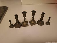 Six variations of small to medium brass candle holders, arranged in a row on a flat surface.