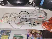 Multiple extension cords and power strips tangled and stacked on shelf