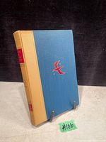 Front view of one hardcover book with beige spine and blue textured cover with red bird emblem on front