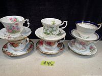 Photo showing six vintage teacups and matching saucers with floral and ornate designs arranged in two rows.