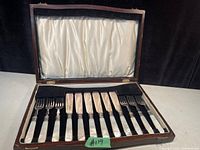 Open wooden box showing 6 dessert forks and 6 dessert knives with mother of pearl handles and sterling silver blades arranged neatly.