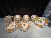 Set of 12 plates stacked with 8 teacups and saucers underneath, showing floral design and gold trim on orange border.