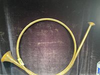 Photo of a vintage solid brass French horn against a dark background, showing its circular body, narrow tubing, and bell flare.