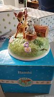 Front-side view of the music box showing Bambi and Thumper figurines on a grass and flower decorated base with original box beneath.