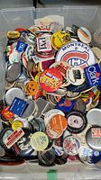 Large plastic bin filled with assorted souvenir buttons showing a variety of political, sports, event, travel, and advertising buttons piled together.