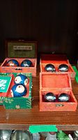 Four sets of two balls in colorful decorative boxes shown on a shelf, two sets painted with dragons, two are metallic silver.