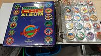 Closed binder labeled 'Collection Cap Album Slammer Whammers Collector Cap Game' with colorful printed caps on front cover and transparent plastic sleeves inside showing round collectible caps.
