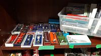 Photo showing a collection of collector spoons in various packaged display boxes arranged on a shelf, including blue, red, green, and white colored backs with spoons inside clear plastic cases.