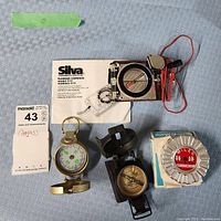 Top view of all four items including Silva Ranger compass, engineer directional compass, black compass, Walk-A-Matic tool, and instruction manual on fabric surface.