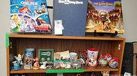 Three Disney-themed hardback books on top shelf: Disney on Ice 2009, Walt Disney World guidebook, The Lion King book. Below, multiple Disney figurines including Tinker Bell, Gus and Jaq mice, Dumbo elephant, and Disney-themed Christmas ornaments and pins.