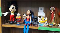 Photo showing a variety of figurines on a wood shelf including Mickey Mouse, robot, Wonder Woman, Sherlock Holmes bust, Yellow Submarine models, and Spanish dancer figurines