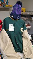 Green Bay Packers varsity jacket with cream leather sleeves and yellow Nike swoosh, displayed with a Dallas Cowboys pom-pom beanie on top.