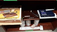 Two Supermatic II cigarette makers placed on a shelf with original packaging boxes. One device is brown, the other blue, both with metal cranks for operation. The packaging shows the product name and cigarette tubes.