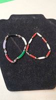 Two sterling silver bracelets on black display stand. One with multi-colored cylindrical spacers and one with alternating red and white rectangular spacers.