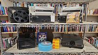 Two white Eumig Mark S 802 Super Single projectors, one black Bell & Howell 851SS, one black Anscomatic 680, film reels, Kodak slide cover, and film boxes on metal shelf in a store