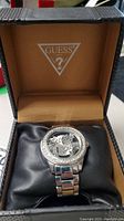 Guess men's watch inside a tan cushioned Guess branded box, showing round face with Guess logo and metal link band with crystal bezel.