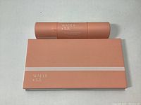 Closed peach-colored makeup palette and matching peach-colored cylindrical makeup stick side by side on white background