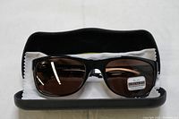 Sunglasses resting inside open case on white cloth, showing the brown polarized photochromic glass lenses and glossy black rectangular frames.