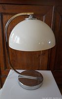 Front view of vintage mushroom-shaped white plastic shade lamp with curved chrome metal arm and round chrome base on white surface with wood background.