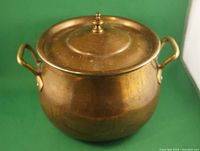 Front side view of the vintage pot with lid on a green background, showing the brass handles and patinated body