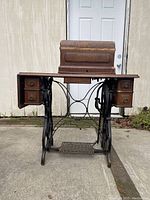 Front view of antique Standard treadle sewing machine in wooden cabinet with drawers and cast iron treadle base in rustic outdoor setting.
