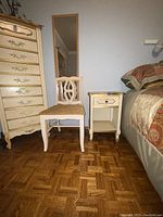 Wood chair with cream and gold upholstered seat next to small nightstand with one drawer and open shelf in cream and gold, both placed on parquet flooring beside a bed.